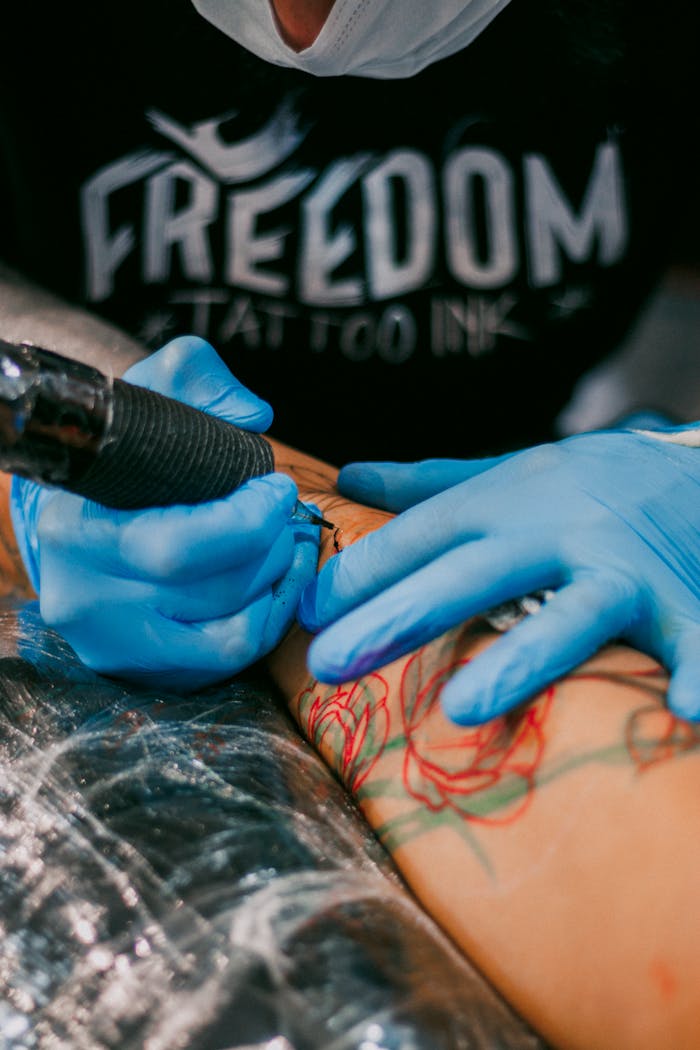 Tattoo artist wearing blue gloves creating a colorful design on a client's arm with 'Freedom Tattoo Ink' in the background.