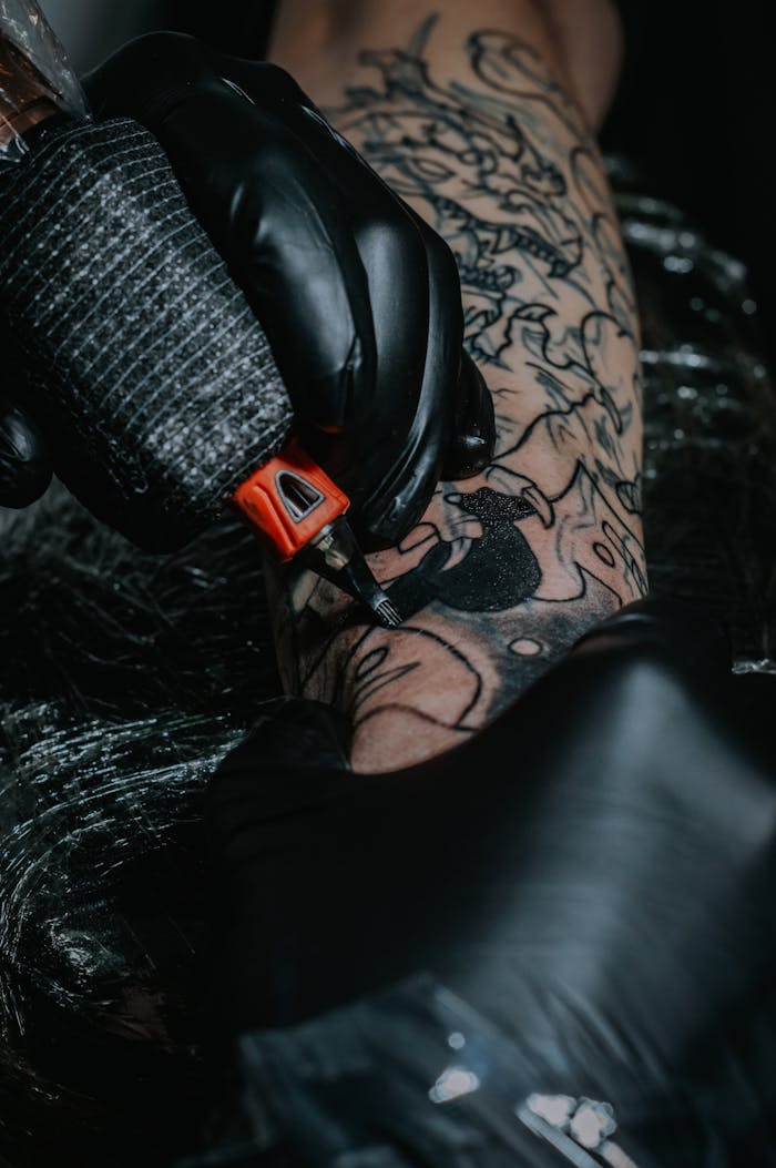 gallery-6 Intricate black tattoo being inked by a gloved artist, showcasing detailed artistry on arm.