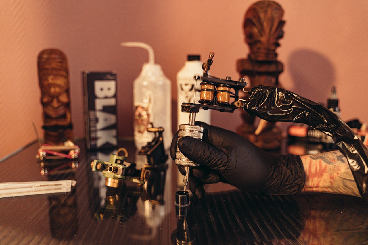 contact-img Tattoo artist's hand preparing tattoo machine in a studio setting with equipment visible on the table.